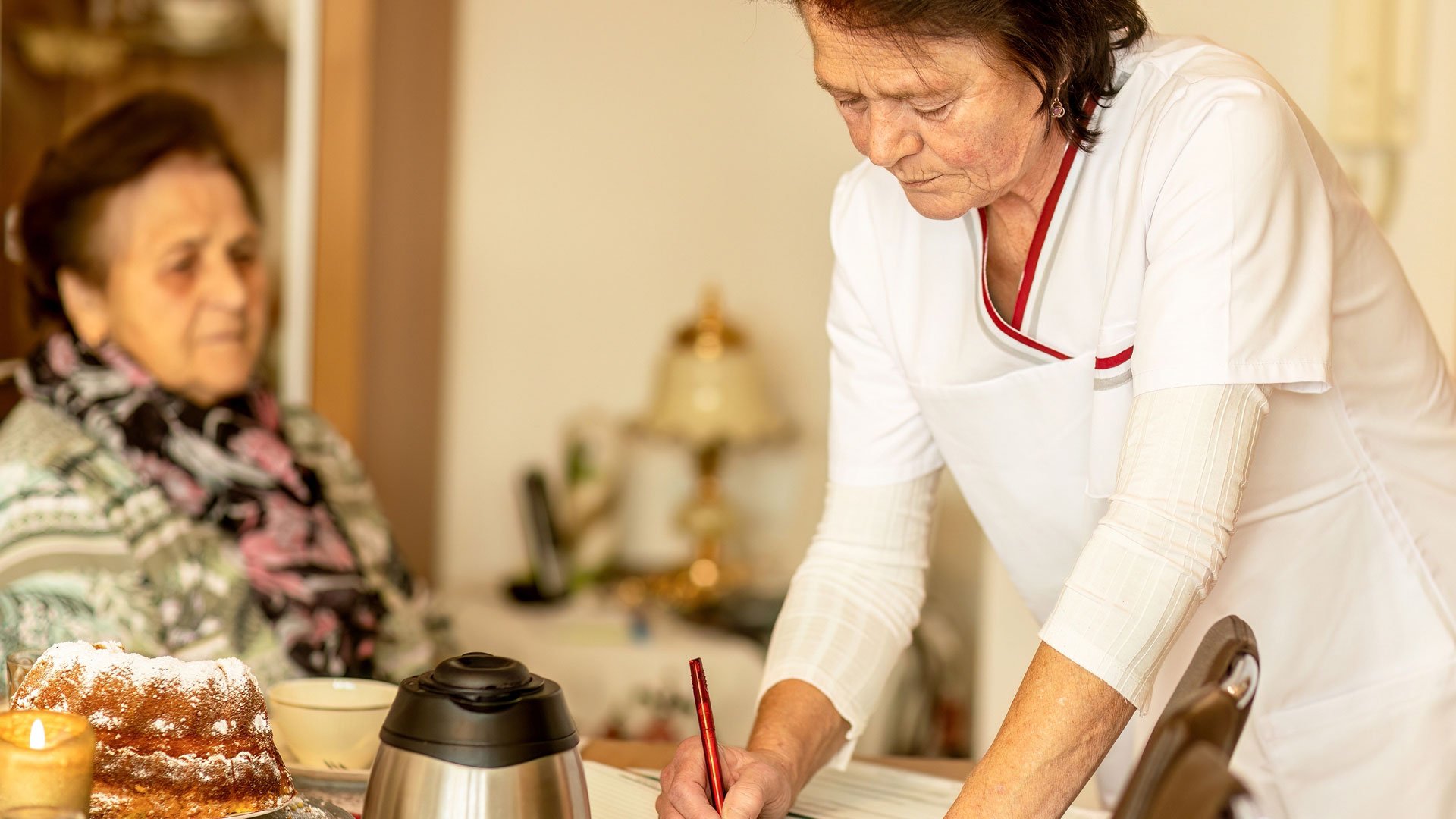 Nurse documents care at a table, resident in background