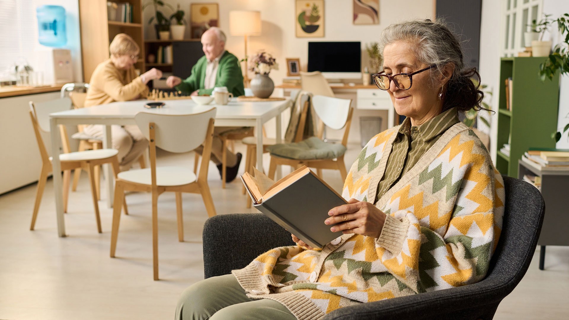 Woman sits in chair reading, other residents at table behind