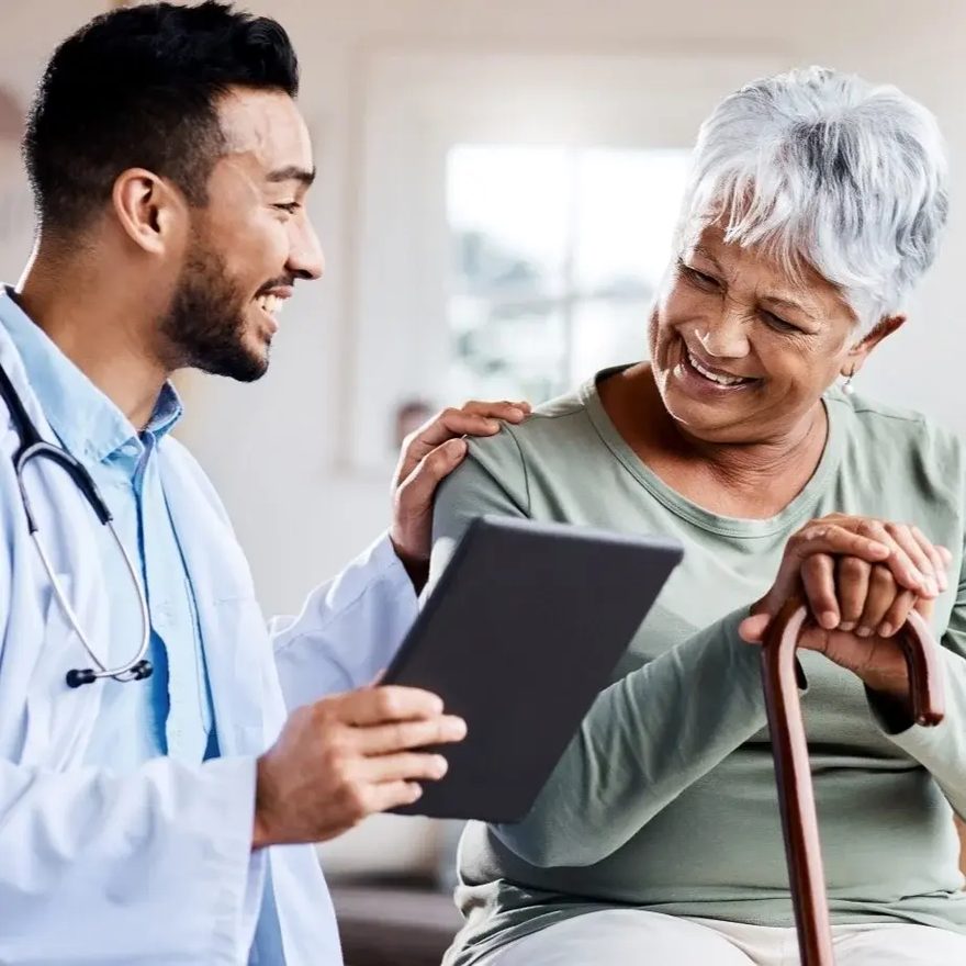 A care worker sitting with an older woman, showing her something on a tablet while they both smile.