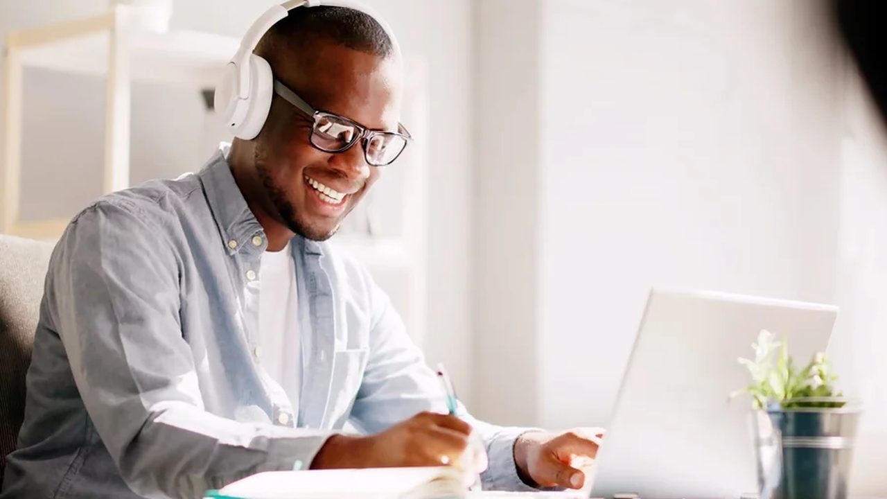 A care worker in headphones taking an online course on a laptop, making notes as he goes.